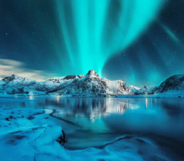 Photo of Aurora borealis over snowy mountains, frozen sea coast, reflection in water at night. Lofoten islands, Norway. Northern lights. Winter landscape with polar lights, ice in water. Starry sky with aurora