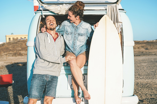 Happy surfers couple standing behind on vintage camper mini van - Young people adventuring on road trip with a minivan transport - Concept of travel, vacation, relationship and youth lifestyle