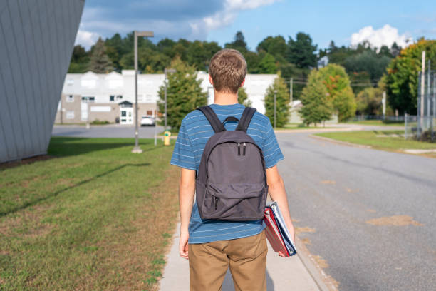 The image displays a male teenager with his back turned to the camera and walking towards a school. He is wearing a backpack and carrying some binders.
