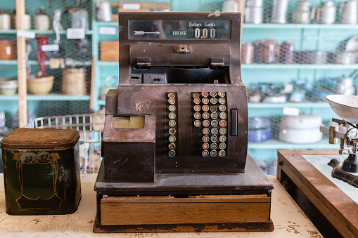 horizontal image of a very old vintage antique wooden cash register with shelves full of dishes in the background.