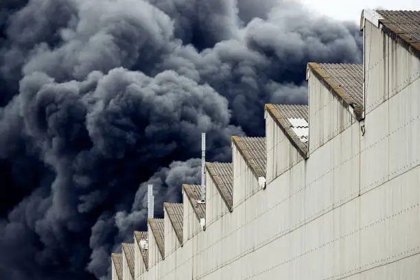 A toxic fire in an industrial district creates plumes of dangerous smoke billowing up into the air as seen from behind a near by warehouse building.