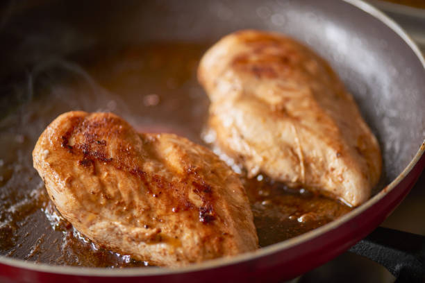 Sizzling in oil are two fresh chicken breasts cooking in a non stick red frying pan.