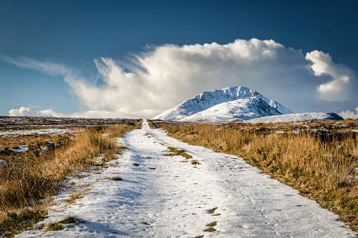 This is an remote dirt road covered in snow that disappears over the horizon.  In the distance a snow capped Errigal mountain can be seen surrounded by white clouds.  This picture was taken in Donegal Ireland in winter.