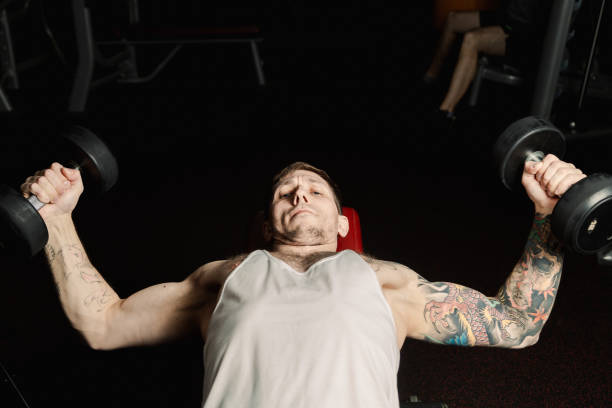 istock Handsome athletic man trains pectoral muscles by breeding dumbbells while lying in the gym 1127246676