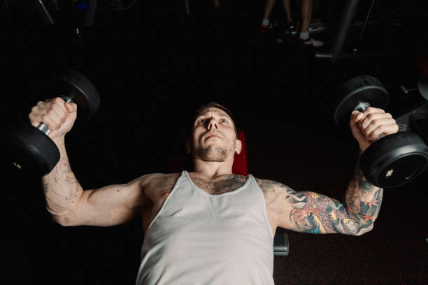 istock Handsome athletic man trains pectoral muscles by breeding dumbbells while lying in the gym 1127246713