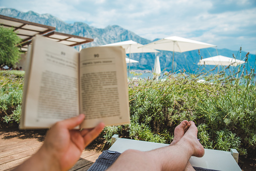 summer vacation. man laying on sun lounger reading book with beautiful view of mountains