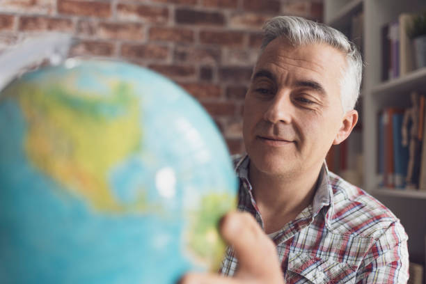 smiling man holding a globe and searching locations, he is planning his vacations and an international travel - senior adult cheerful adventure discovery imagens e fotografias de stock