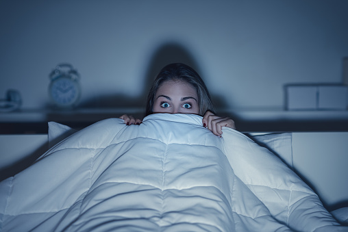 Woman watching a scary horror movie on tv late at night, she is frightened and hiding under the blanket
