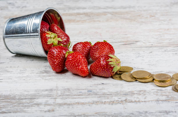Strawberries and coins. Finance A bucket of fresh ripe strawberries and a pile of scattered coins close-up on a light wooden background, copy space. монета stock pictures, royalty-free photos & images