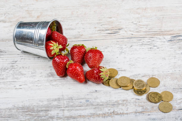 Strawberries and coins. Finance A bucket of fresh ripe strawberries and a pile of scattered coins close-up on a light wooden background, copy space. монета stock pictures, royalty-free photos & images