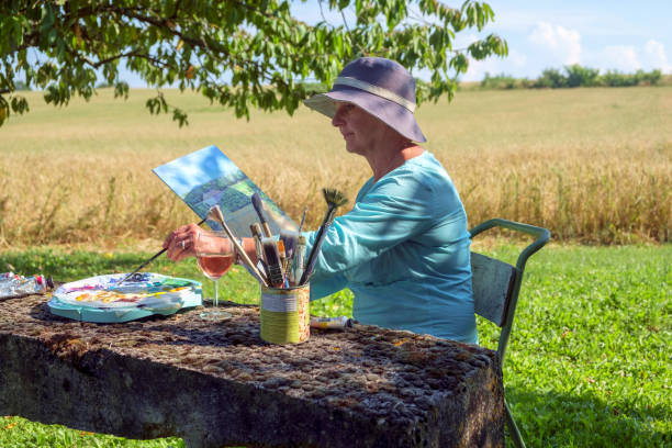 An artist sits with a rural countryside view painting A lady artist sits in late afternoon shade with a rural countryside view working on the painting she started earlier in the day. painted image paintings oil paint senior women stock pictures, royalty-free photos & images