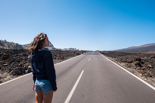 Lonely beautiful young caucasian woman standing in front of a long way straight road in the middle of the lava landscape and mountains. travel and wanderlust experience and concept for lonely female traveler