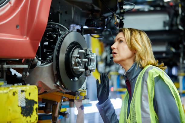 Blond female engineer checking vehicle at factory Confident mature female maintenance engineer examining car at factory. Blond manager is checking vehicle at automobile industry. She is wearing reflective clothing. car plant stock pictures, royalty-free photos & images