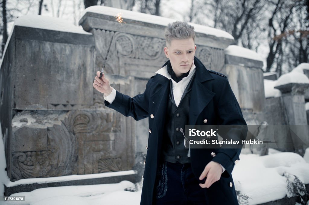 Young man in image of black magician makes a spell using magic wand. Young man in image of black magician makes a spell using magic wand next to crypt. Adult Stock Photo