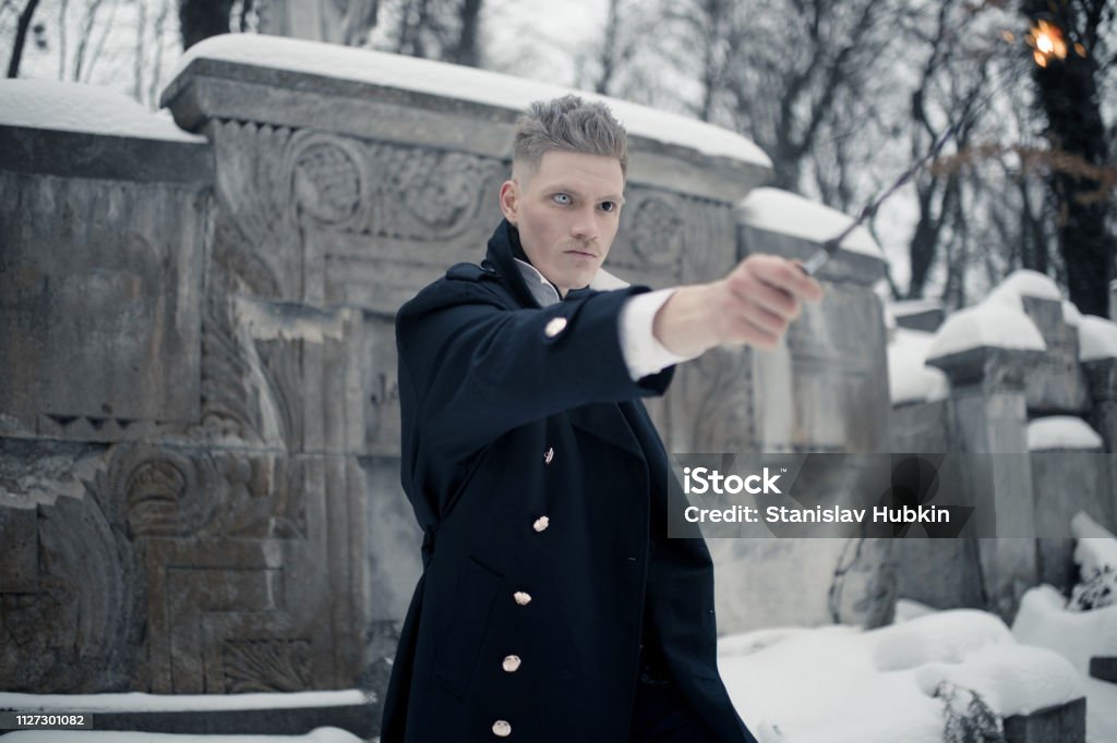 Young man in image of black magician makes a spell using magic wand. Young man in image of black magician makes a spell using magic wand next to crypt. Adult Stock Photo