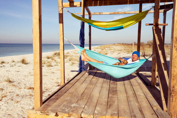 Joyful traveler with funny dog relaxing in a hammock on bungalow veranda next to turquoise sea. Awesome adventure travel to the seashore. Wide angle.