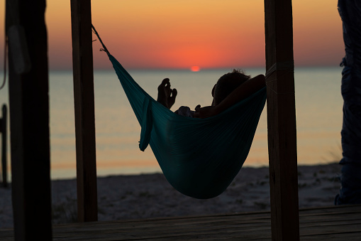 Man relaxes in a hammock on abandoned bungalow veranda, admiring the turquoise sea during sun set. Awesome travel to the seaside. Back view, backlight.