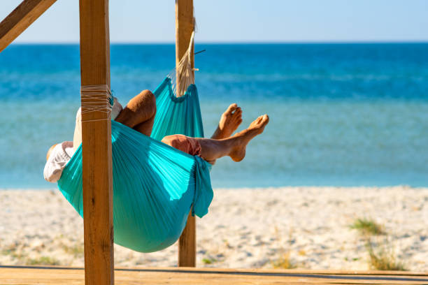 Man relaxes in a hammock on abandoned bungalow veranda, admiring the turquoise sea in front of him. Amazing sunny day on the beach. Back view.