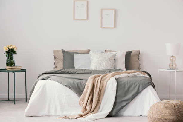 istock Flowers on table next to bed with pillows in white bedroom interior with posters and pouf. Real photo 1127303247