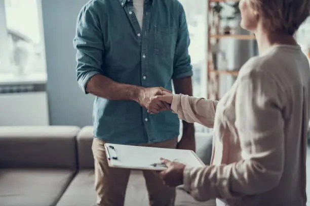 Man in casual clothes standing opposite his reliable psychotherapist and shaking her hand while coming to the meeting