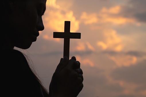 Silhouette of woman hand holding holy lift of christian cross with light sunset background.
