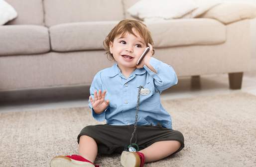 Cute baby boy talking with imaginary friend on cellphone, playing on floor at home, copy space