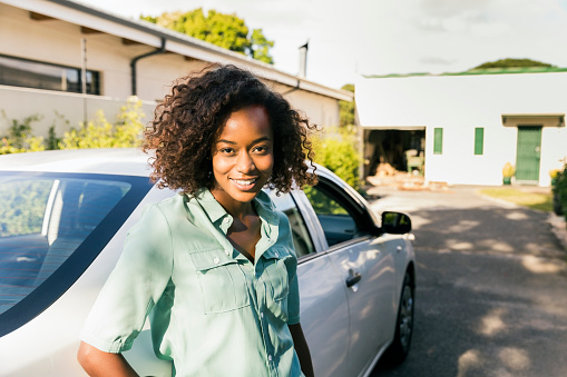 Portrait of happy woman standing by car on street. Confident young female is outside house on sunny day. She is in casuals.