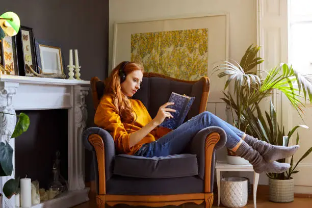 Full length of young woman reading book while listening to music. Side view female is resting on chair at home. She is wearing casuals.