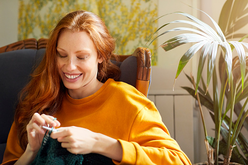 Woman knitting while sitting by plant on chair. She is smiling at home. Young female is wearing casuals.