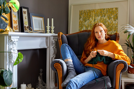 Portrait of woman knitting while sitting on chair. Confident female is by fireplace at home. She is wearing casuals.