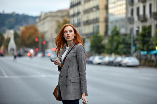 Beautiful redhead woman looking away in city. Young female commuter is walking on city street. She is wearing gray blazer.