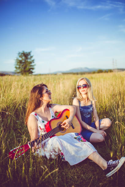 Two young girlfriends sitting on the meadow and relaxing with guitar.