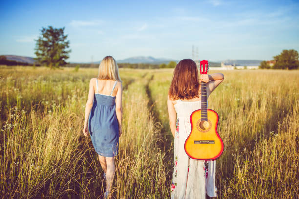 Two caucasian woman walking through the meadow with guitar.