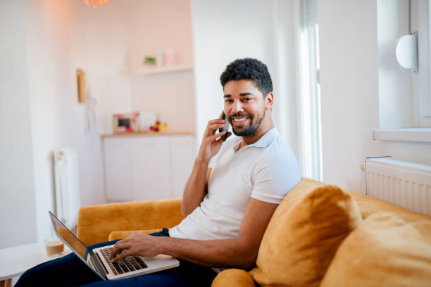 istock Handsome mixed-race man talking on smart phone and using laptop at home, looking at camera. 1127360312