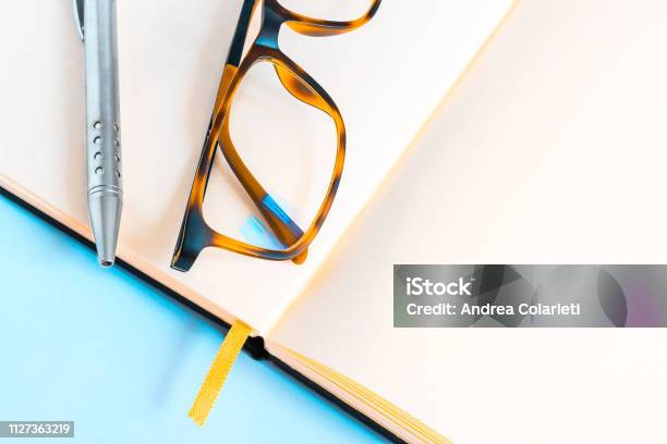 A Pair Of Elegant Eyeglasses And A Ballpoint Pen On An Open Notebook On A Blue Background Stock Photo - Download Image Now