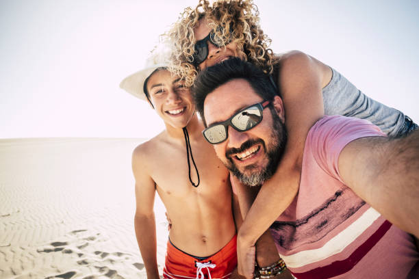 szczęśliwa rodzina na świeżym powietrzu na plaży podczas wakacji. turyści robiący sobie selfie w szczęściu razem w wakacje z piaszczystymi wydmami uciekają w tle. ciesz się wolnością i stylem życia z uśmiechami i młodzieńczym śmiechem - men footprint beach sunset zdjęcia i obrazy z banku zdjęć