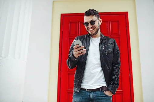 Image of handsome male in black leather jacket reading a messeage from a cellphone and smiling over a red door