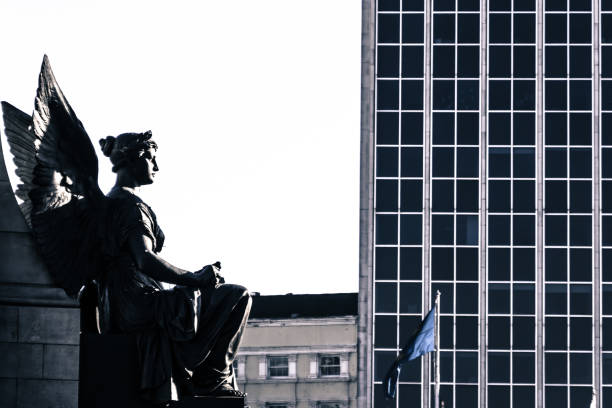 Dublin, Ireland - February 2, 2019: Profile of a winged figure on the O'Connell monument looking upstream along the River Liffey towards the cityscape beyond.  The O'Connell monument is at the foot of O'Connell Bridge in Dublin's city centre.  It celebrates the Irish lawyer and politician, Daniel O'Connell (1775-1847), who struggled for the emancipation of Irish Catholics and self-determination for the people of the island of Ireland.  The statue is the work of John Foley and was completed in 1882.  The modern office building (rhs) is O'Connell Bridge House (1965), designed by Desmond Fitzgerald.