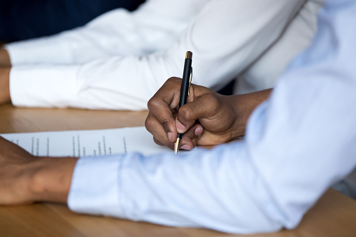 Businessman sitting at desk holds pen putting signature on official paper signing document, close up focus on hand. Human resources, good deal or agreement between an insurance company and the insured