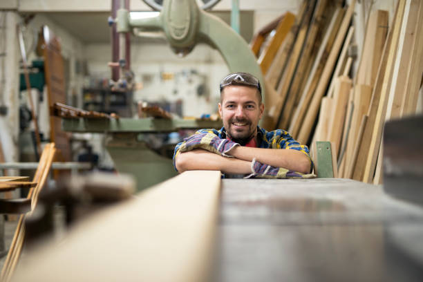 ritratto di giovane falegname sorridente in piedi accanto a una macchina e materiale legnoso nel suo laboratorio di falegnameria. - work tool carpentry construction wood foto e immagini stock