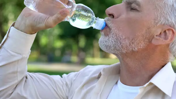 Mature man drinking water from bottle in park, maintaining water balance