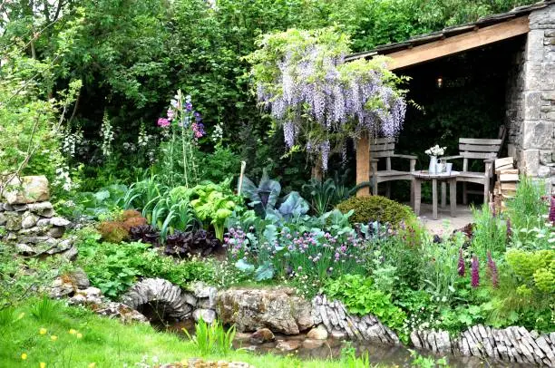 A vegetable garden intermingled with soft planting of sweet peas, foxgloves and wisteria growing next to a stone walled stream within a show garden on Main Avenue at Royal Horticultural Society Chelsea Flower Show 2018