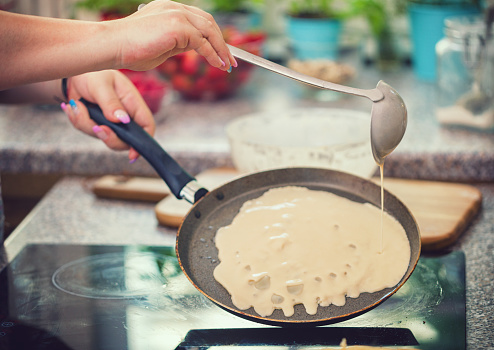 Young woman making crepes at home, she pouring dough into frying pan