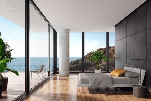 Modern luxurious bedroom in a seaside villa with black stone wall Luxurious minimalist bedroom in a seaside villa with big panoramic windows
and ocean view. White textile king size bed with white metal construction in front of 
black wall with natural stone large tiles. Round modern gray nightstand with a
modern metal lamp next to the bed. Wooden varnished floor. Big terrace with an
ocean view and glass fence with green plants. Summer daylight scene.
Background is my own photo. building terrace photos stock pictures, royalty-free photos & images