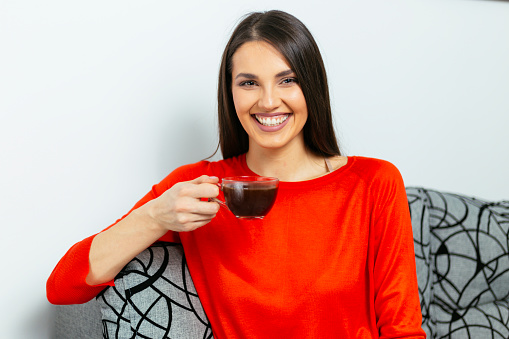 Portrait of beautiful woman sitting on sofa and holding cup of coffee while smiling