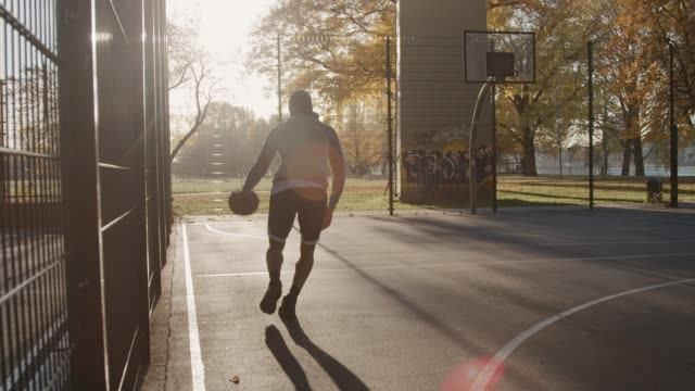 Slow motion shot of sportsperson dribbling basketball. Male athlete is scoring in hoop. He is playing on outdoor court during sunny day.