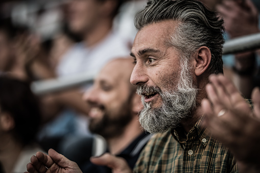 Portrait of a middle-aged man with gray hair and beard watching a sports game in a stadium.