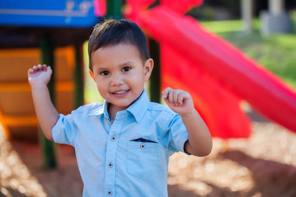 A toddler boy with arms raised up and a smile on his face, playing in a colorful kids playground. stock photo