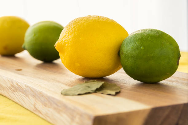 A whole lemon is isolated among other fresh fruit. The fruit are all set on a wooden cutting board and yellow table top with a white background.