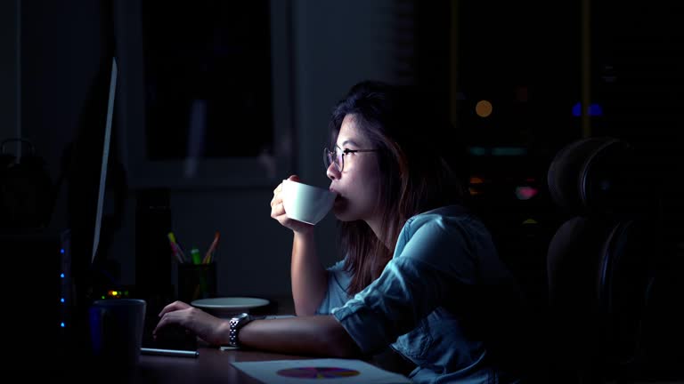 4k footage scene of Attractive Asian woman working late and drinking coffee with serious action on the table in front of computer monitor desktop at workplace in the dark, Work late and Work hard concept